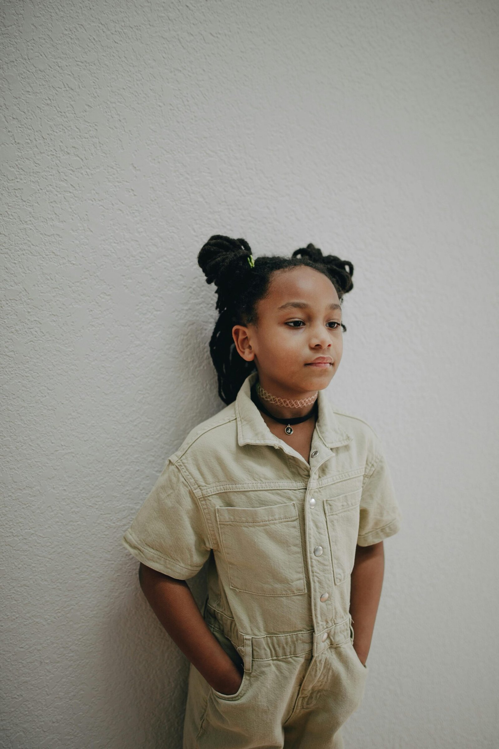 A young girl poses indoors in overalls, showcasing her Afro pigtails and confident expression.