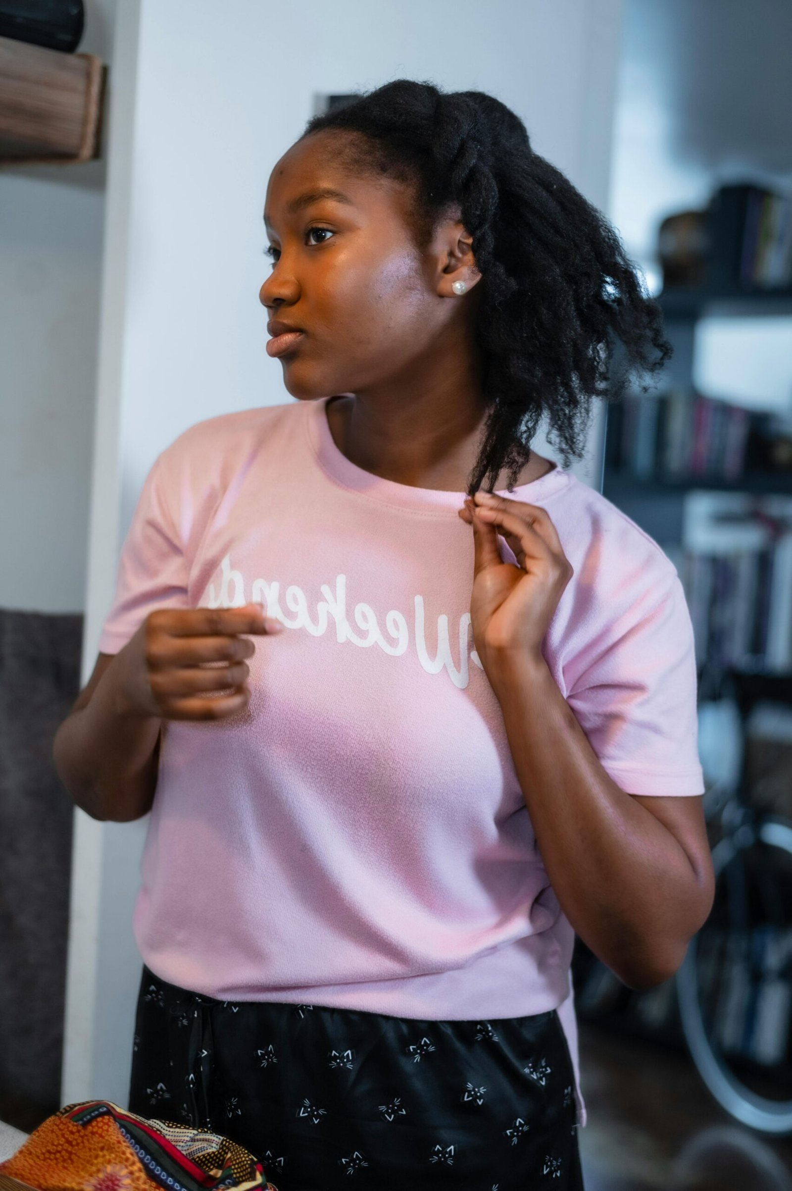 Young African woman with curly hair adjusting her look indoors, wearing a pink sweater.